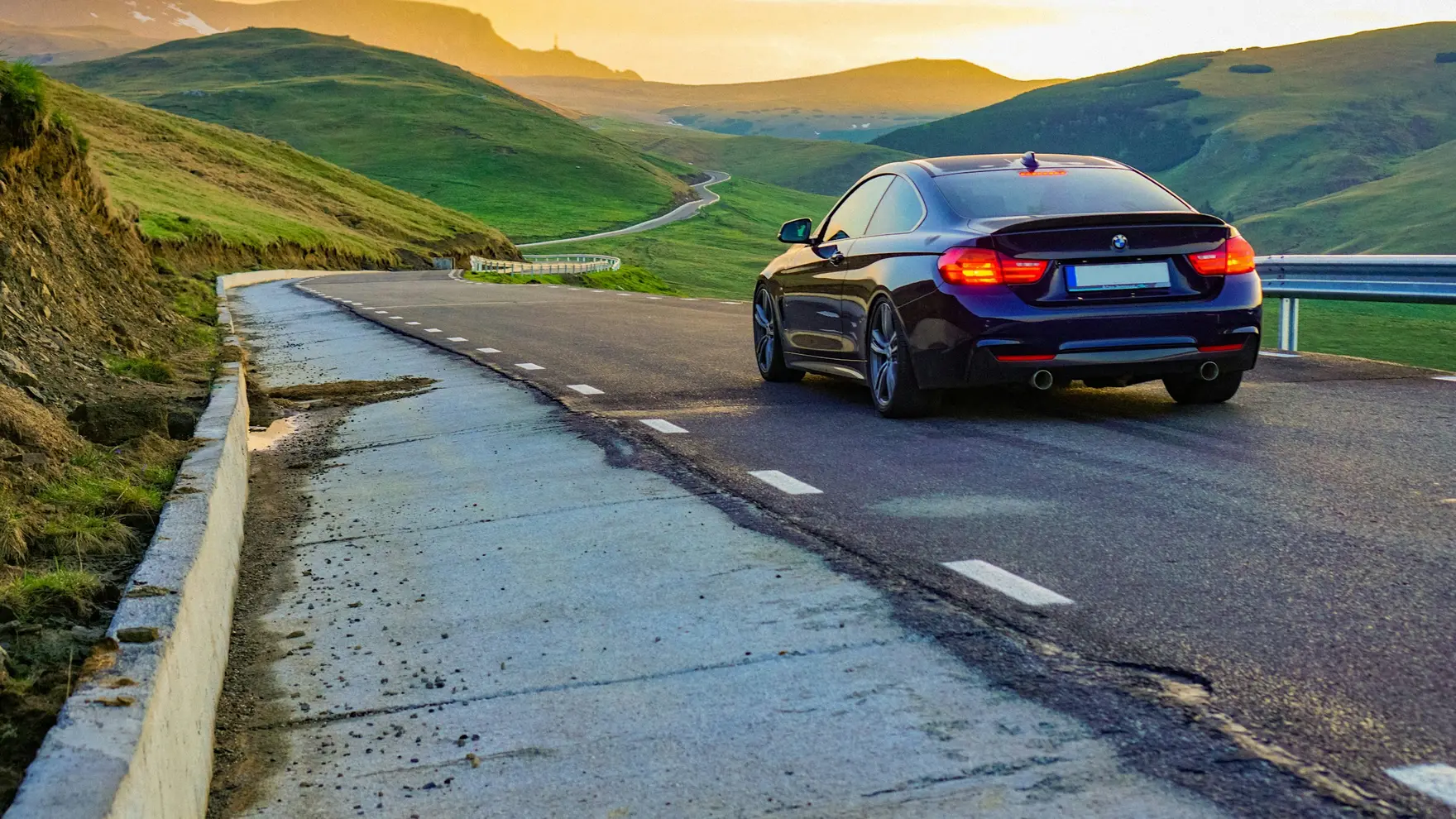 Dunkles Coupé fährt bei Sonnenuntergang über eine Passstraße Richtung Horizont